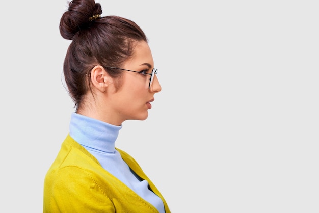 Photo side view portrait of young brunette woman has serious facial expression wears transparent eyewear and casual outfit looking to blank copy space isolated over white background