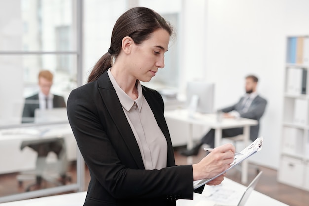 Side view portrait of young businesswoman writing on clipboard while standing in office, copy space