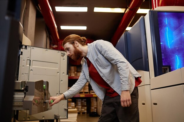 Side view portrait of young man doing maintenance check on\
industrial printing machines in shop