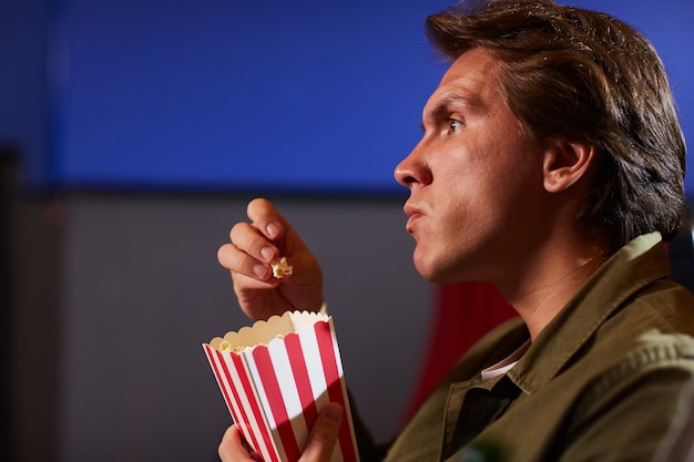 Side view portrait of young man eating popcorn while watching movie in cinema theater alone, copy space