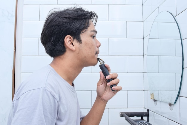 Side view portrait of young man shaving with an electric razor in bathroom mirror at home