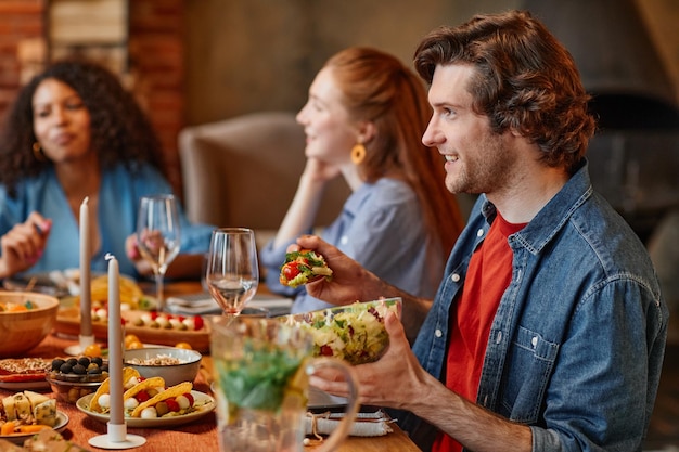 Side view portrait of young man talking to friends at table during dinner party in cozy setting copy