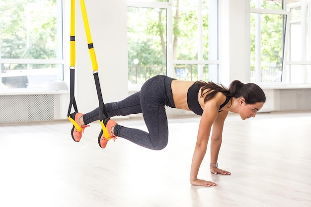 Side view portrait of young muscular woman wearing black sportswear standing on plank position use fitness straps doing push ups while legs hanging on trx Indoor window background abs exercises