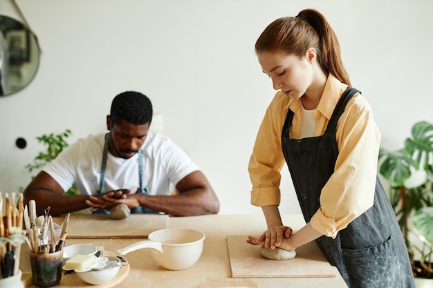 Photo side view portrait of young woman shaping clay and demonstrating technique to people in pottery stud