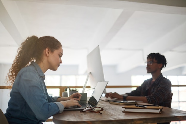 Photo side view portrait of young woman using laptop while working at desk in software development agency with african-american colleague writing code in background, copy space