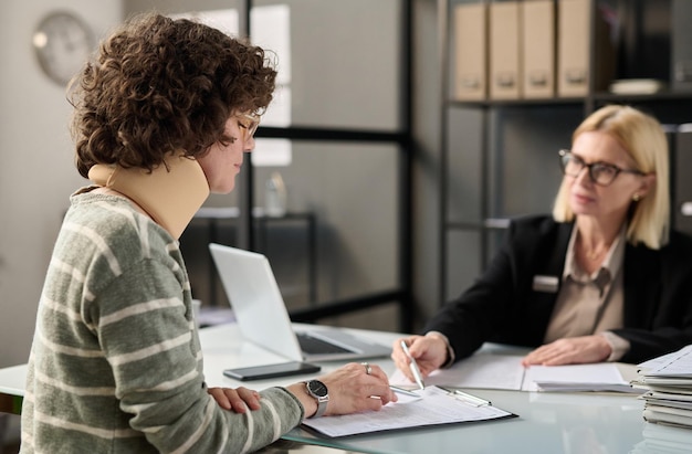 Side view portrait of young woman with neck injury filling documents at insurance agency office copy