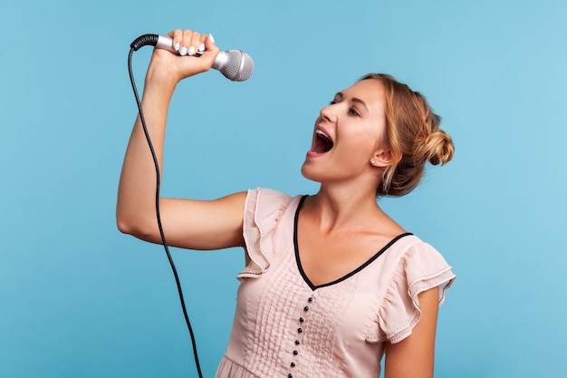 side view positive blonde woman in summer dress loudly singing song, holding microphone in hand, having fun resting in karaoke, singer. indoor studio shot isolated on blue background.