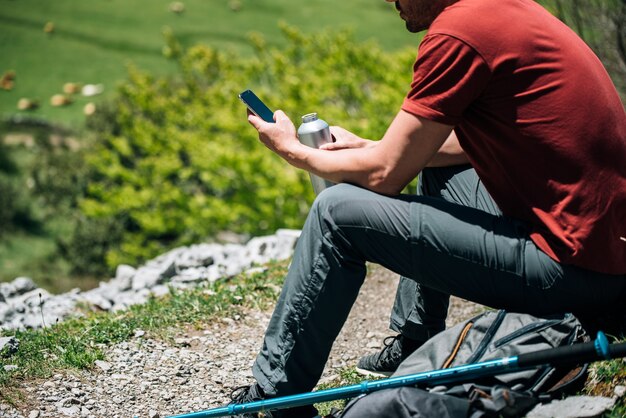 Side view of positive hiker sitting on backpack and scrolling mobile phone while resting with water bottle in rocky terrain