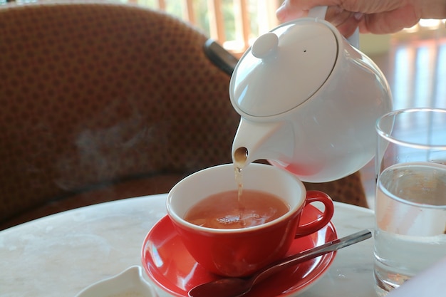 Photo side view of pouring tea with white tea pot and red tea cup in the time of tea break