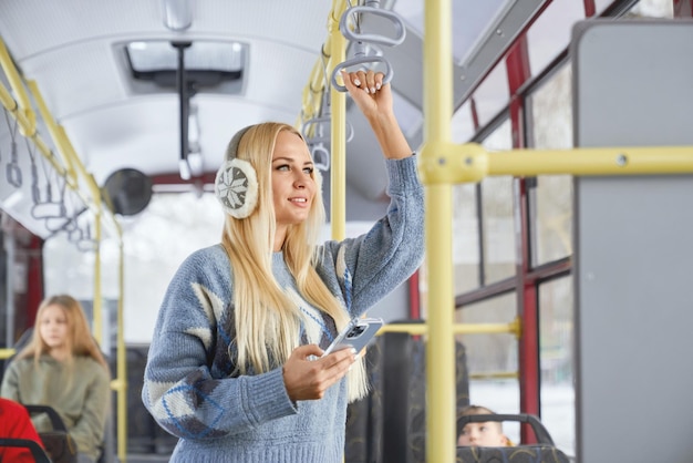 Side view of pretty blonde girl standing with mobile looking at window smiling in public transport