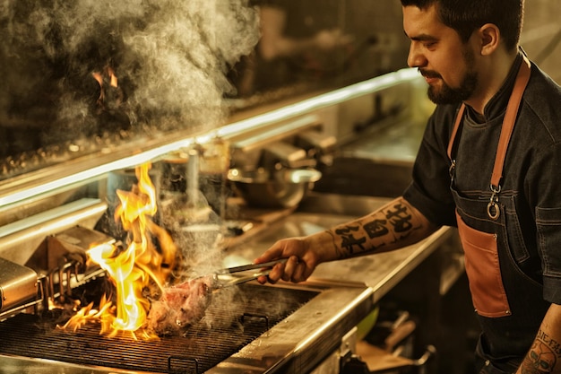 Side view of professional chef cooking delicious juicy beef\
steak on flaming grill handsome man with beard and tattoos on hand\
preparing food in modern restaurant kitchen