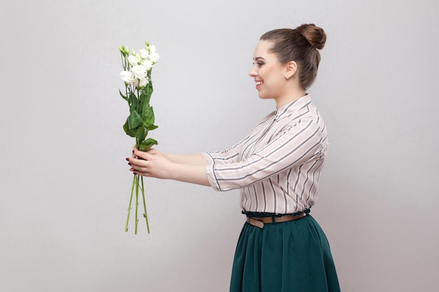 Side view profile of attractive romantic young woman in striped shirt and green skirt holding bouquet of white flowers and giving you. Indoor, studio shot, isolated on gray background, copy space