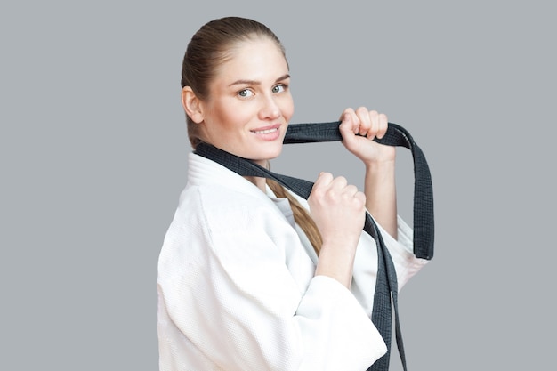 Side view profile of happy beautiful young athletic woman in white kimono with black belt wraps around the neck standing, holding belt and toothy smiling. Indoor studio shot, isolated, grey background