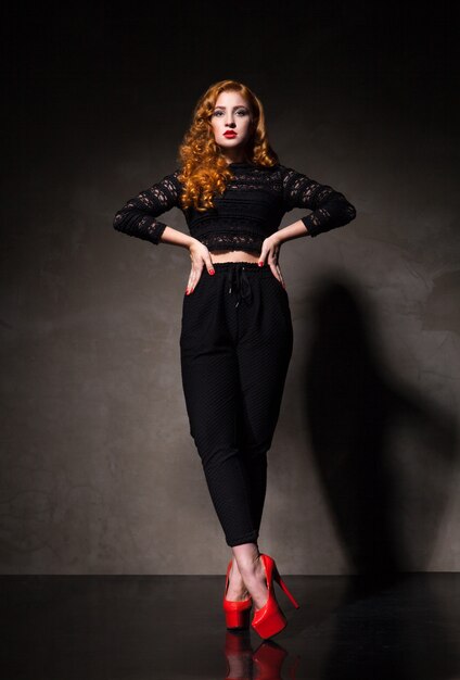 Side view of red-haired woman in black and red high heels posing against of black wall