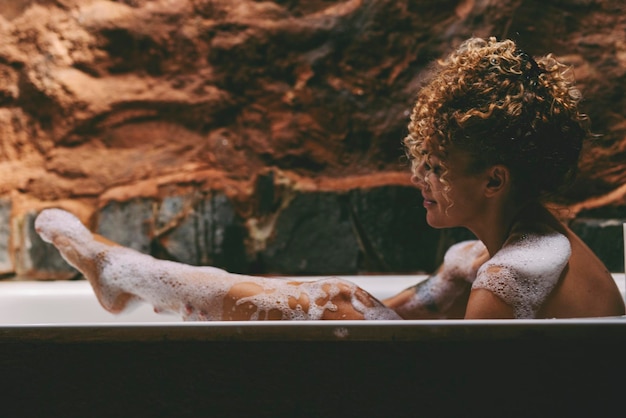 Photo side view of relaxed african american naked young woman taking bath at home laying in beautiful bathtub rubbing her skin with sponge and smiling copy space home spa hygiene relaxation concept