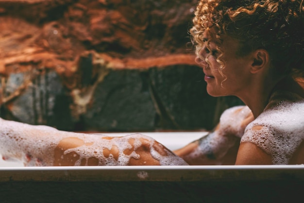 Side view of relaxed african american naked young woman taking bath at home laying in beautiful bathtub rubbing her skin with sponge and smiling copy space Home spa hygiene relaxation concept