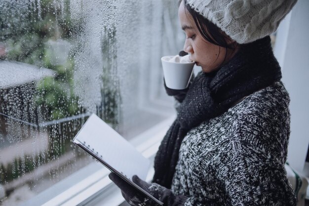 Боковой вид расслабленной женщины, пьющей кофе во время чтения книги и стоящей возле окна в дождливый день