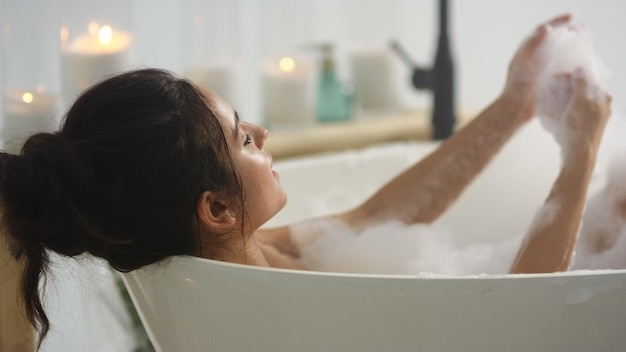 Side view of relaxed woman touching skin with foam in bath Close up sexy girl taking bathtub at home Romantic brunette woman washing hands in bath in slow motion