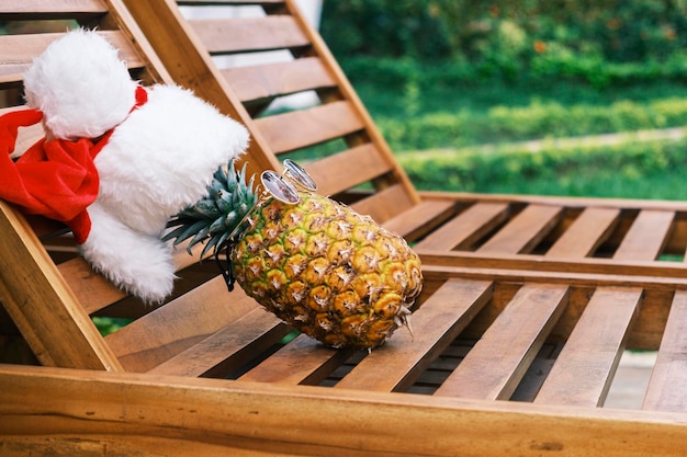 Вид сбоку на расслабляющий забавный ананас в солнцезащитных очках и шляпе санта-клауса лежит на деревянном солярии