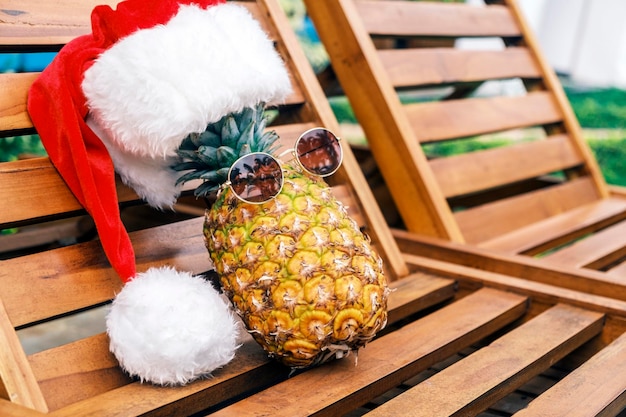 Photo side view of relaxing funny pineapple wearing sunglasses and santa claus hat lies on wooden sun loun