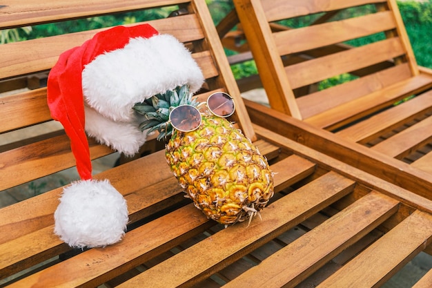 Side view of relaxing funny pineapple wearing sunglasses and santa claus hat lies on wooden sunbed i