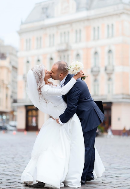 Vista laterale della sposa e dello sposo romantici che indossano abiti alla moda che si abbracciano e si baciano mentre posano sulla strada della città antica durante la passeggiata di nozze