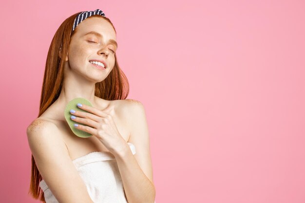 Side view of satisfied female model with clean freckled skin, long red hair, wiping body with shower sponge, enjoying closed eyes, cute smiling isolated over pink background.