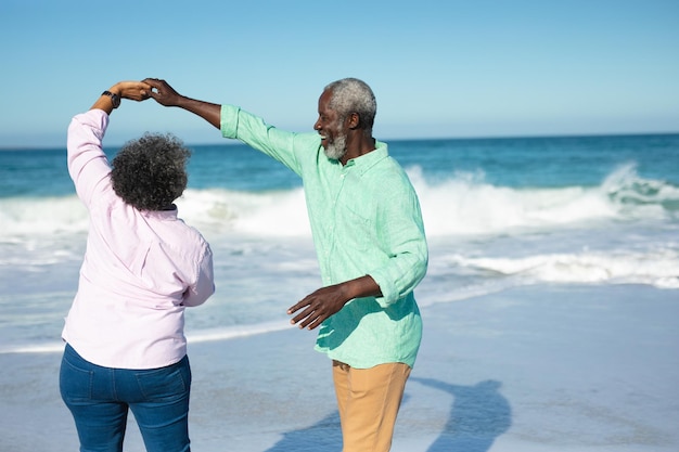 青い空と海を背景にビーチに立ち、手をつないで踊り、楽しんでいるアフリカ系アメリカ人の年配のカップルの側面図