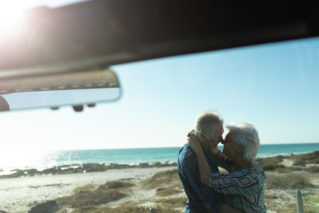 太陽の下のビーチで立って抱きしめ合っている高齢の白人カップルの横の景色車の窓から見るとお互いに見つめ合い笑顔です