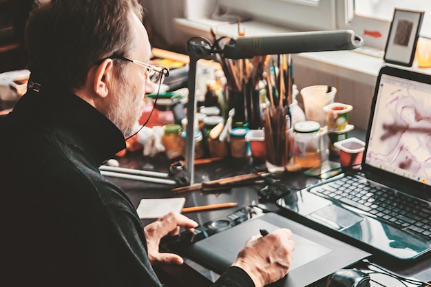 Photo side view of senior man using laptop and graphic tablet on desk