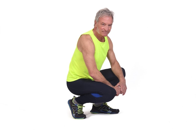 Side view of a senior man with sportswear squatting looking at camera on white background