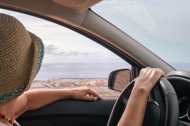 夏の帽子をかぶった年配の女性が車の中に座って、ハンドル越しに海の景色を見ている側面図