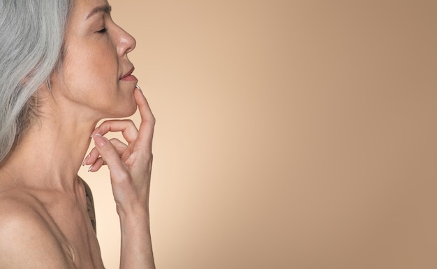 Photo side view of sensual senior goodlooking lady touching her chin posing on beige background free space