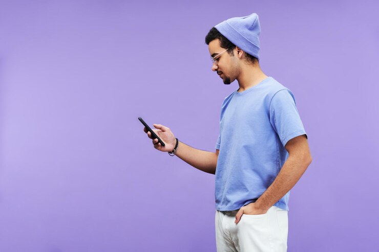 Photo side view of serious guy in blue tshirt and beanie hat using mobile phone