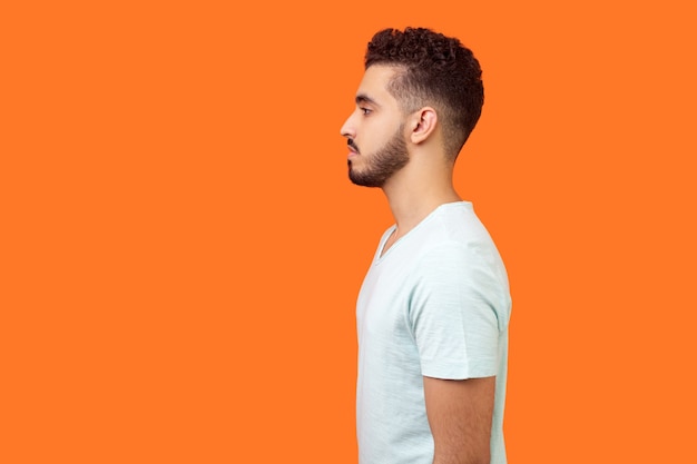 Side view of serious handsome brunette man with beard in casual white t-shirt standing with hands down and looking left, empty copy space for text. indoor studio shot isolated on orange background