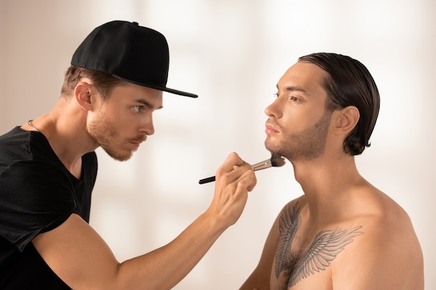 Side view of serious young makeup artist with brush applying powder on chin of male fashion model with tattoo on chest against white wall