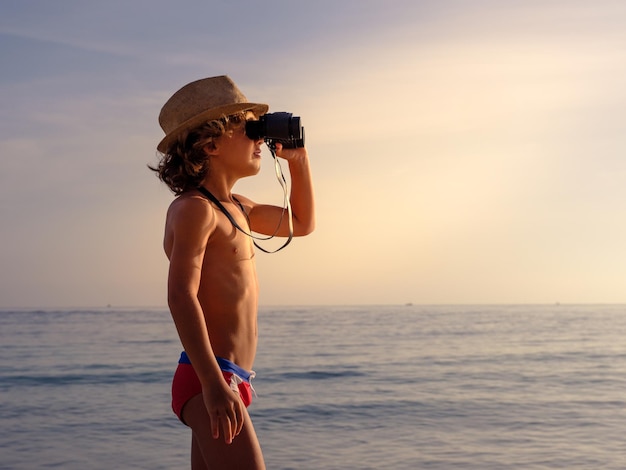 Side view of shirtless boy in straw hat and swimwear observing seascape through binoculars at sundown in tropical resort in evening