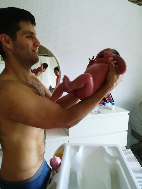 Side view of shirtless father holding newborn daughter crying in bathroom