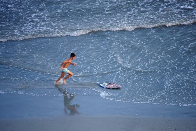 Photo side view of shirtless man running towards skimboard on shore at beach
