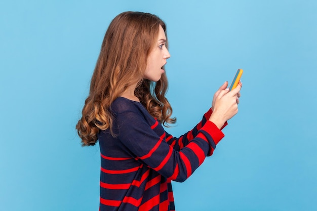 Side view of shocked woman in striped casual style sweater holding smart phone in hands looking at display with astonished expression braking news Indoor studio shot isolated on blue background