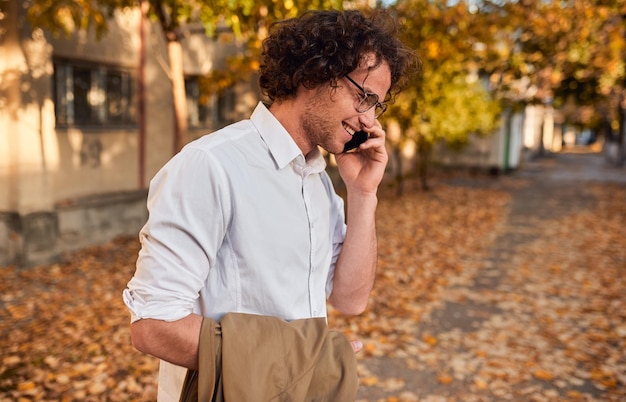 屋外を歩いている間、秋の通りでポーズをとって、彼の携帯電話で話している眼鏡をかけている巻き毛の笑顔の男を呼び出すためにスマートフォンを使用してハンサムなビジネスマンの側面図
