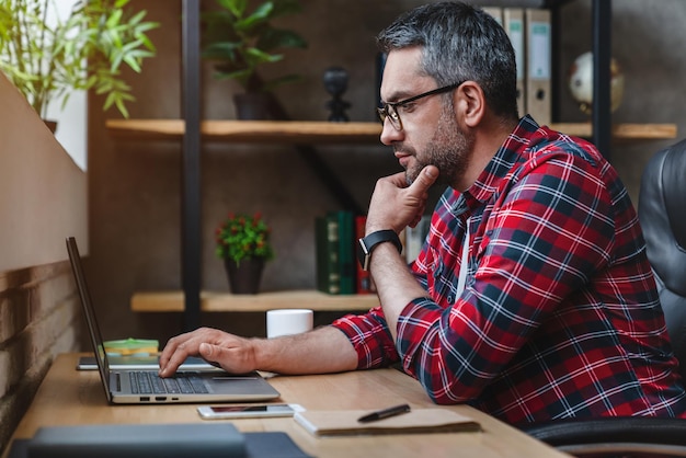 Боковой снимок задумчивого молодого человека, сидящего в домашнем офисе и работающего на ноутбуке