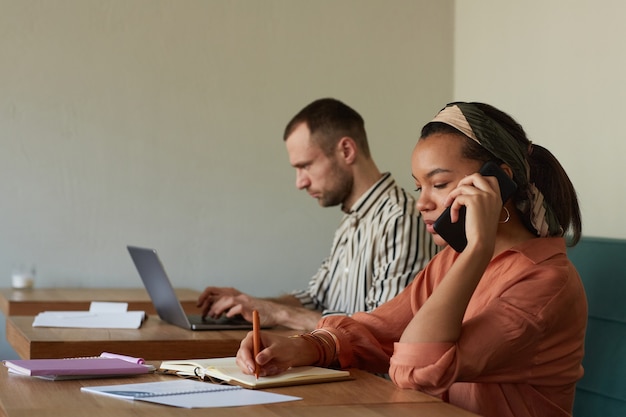 Side view shot of two people working at tables in cafe focus on young African-American businesswoman speaking by smartphone, copy space