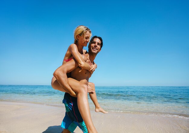 Side view shot of young man caring his girlfriend on back at the beach couple enjoying piggybacking on the sea shore