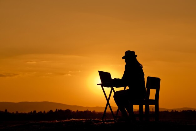 Side view of silhouette young woman using laptop on table while sitting against sky during sunset