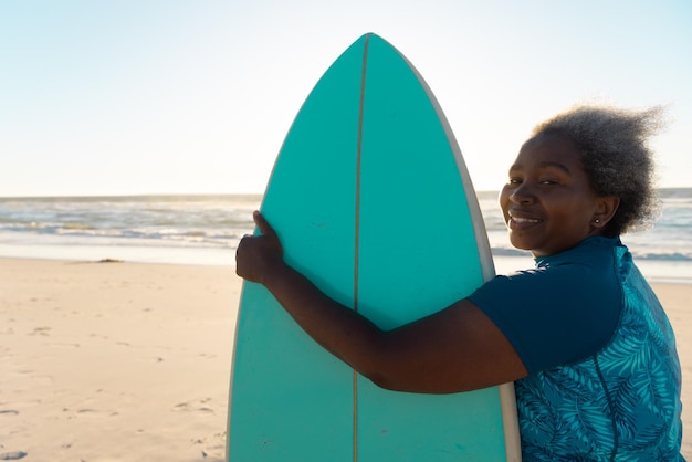 Боковой вид улыбающейся афроамериканской пожилой женщины с доской для серфинга, стоящей на пляже над морем и небом.
