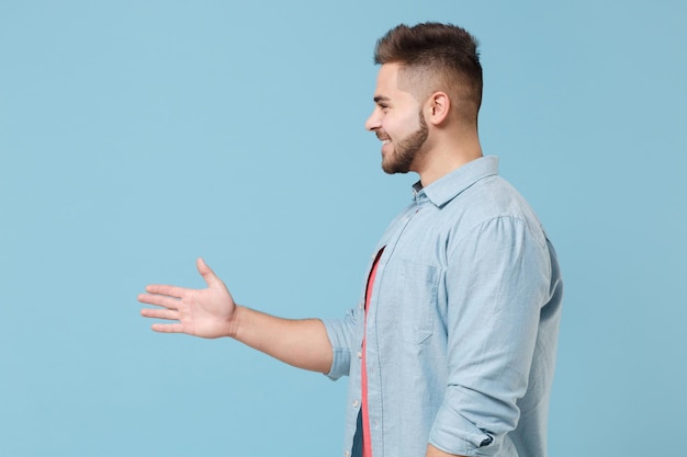 Side view of smiling bearded young guy 20s in casual shirt posing isolated on pastel blue wall background. People lifestyle concept. Mock up copy space. Standing with outstretched hand for greeting.