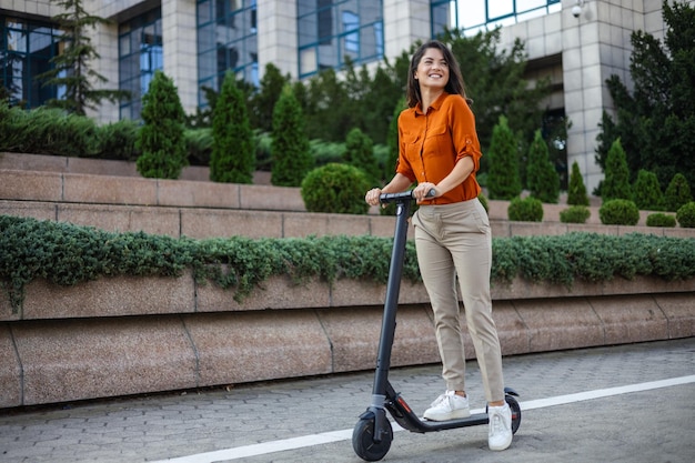 Photo side view of smiling businesswoman commuting to work on the electric scooter near city park on sunny day