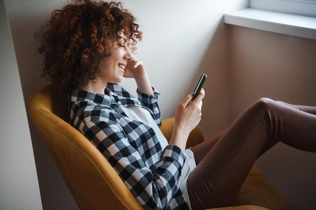 Side view of a smiling curly lady holding a smartphone and checking messages while resting in armcha...