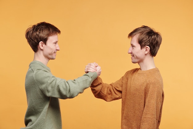 Side view of smiling friendly twins in sweaters standing against orange background and making firm handshake